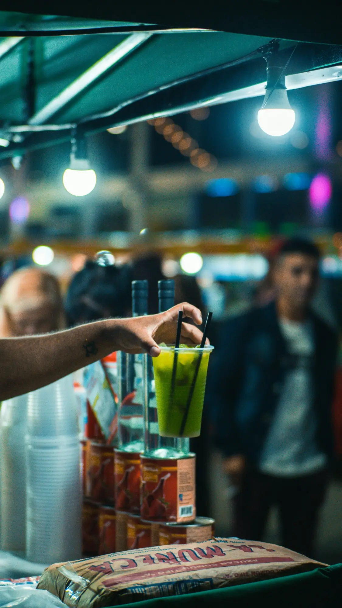 A hand lifting a green iced drink at a night food stall under string lights