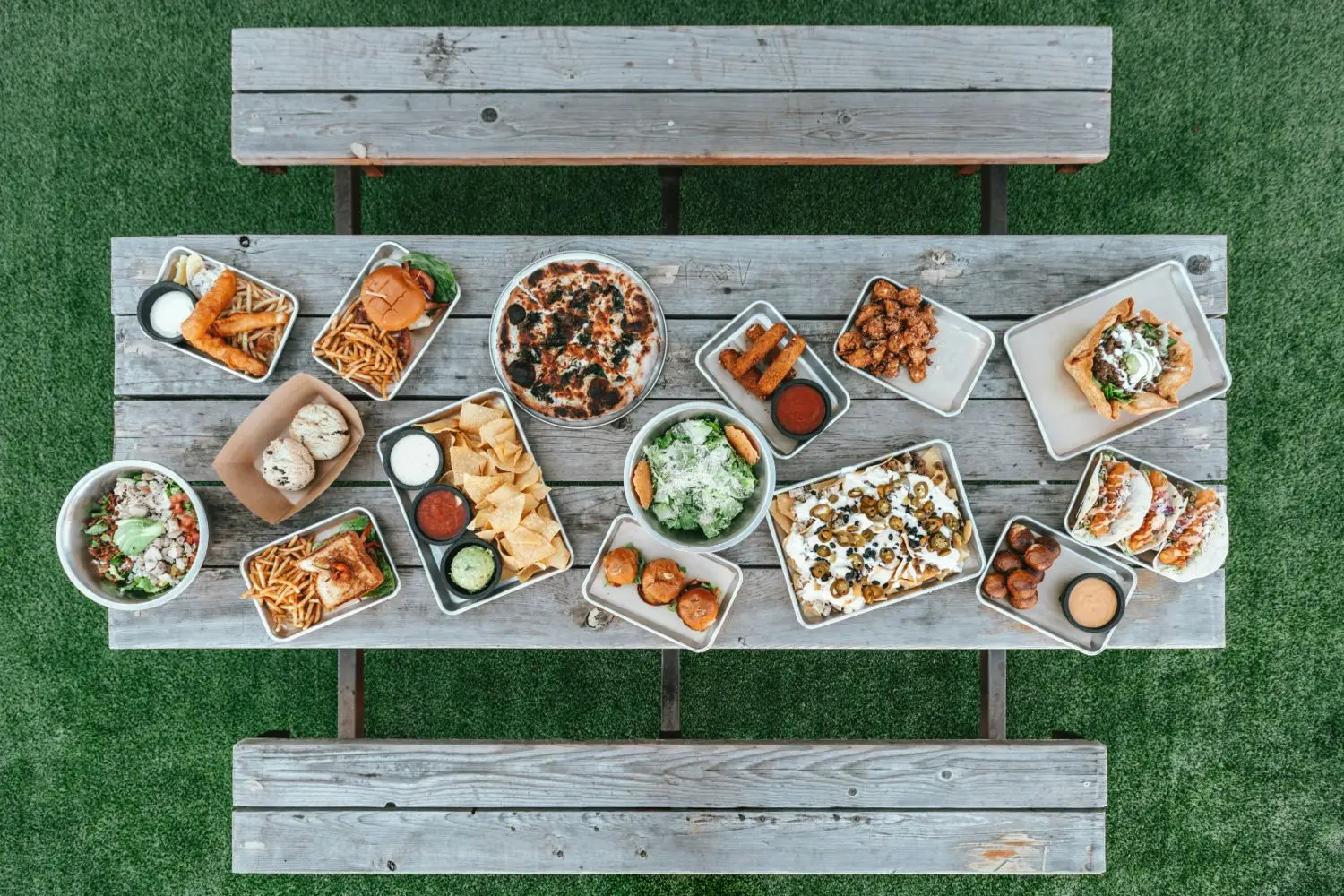 An overhead picnic table spread with burgers, pizza, nachos, salads, and tacos